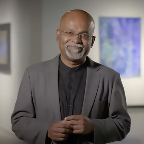 An artist wearing a black shirt and brown sports coat stands in an art gallery. The background of the image is blurred to create a bokeh effect.
